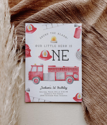 firetruck first birthday party invitation with playful watercolor graphics of firetruck, hose, hat, ladder, hydrant, extinguisher and siren light.  Reads "sound the alarm! Our little hero is ONE".  A fireman's hat is used in place of the "o" in ONE.