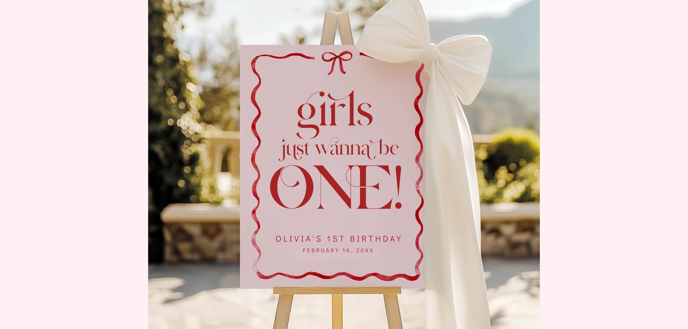 A pink and red sign on a wooden easel that reads 'girls just wanna be ONE! Olivia's 1st Birthday February 10, 2026' with a white bow tied to the top, photographed outdoors with a blurred garden background.