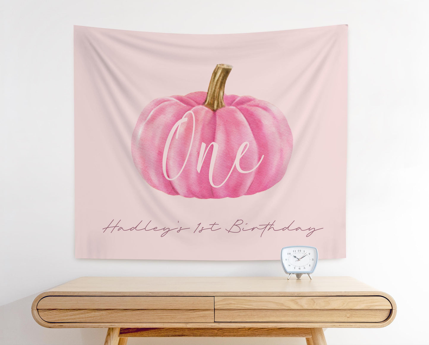 pale pink fabric backdrop with a pink watercolor pumpkin graphic.  "ONE" is written in modern script overlaying the pumpkin.  Below in smaller text reads "Hadley's 1st Birthday".  Clean, Minimal design for girl first birthday party