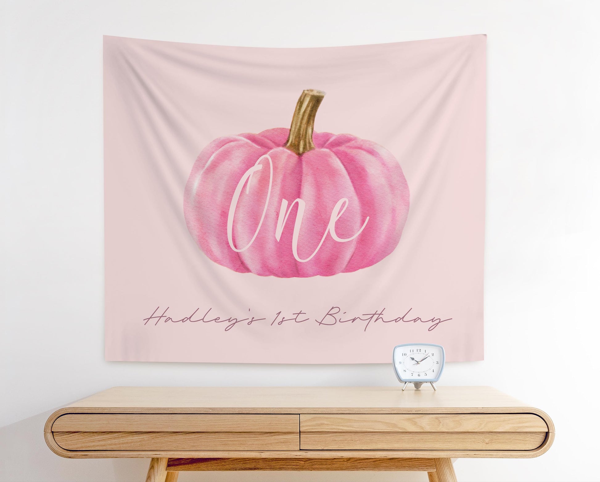 pale pink fabric backdrop with a pink watercolor pumpkin graphic.  "ONE" is written in modern script overlaying the pumpkin.  Below in smaller text reads "Hadley's 1st Birthday".  Clean, Minimal design for girl first birthday party