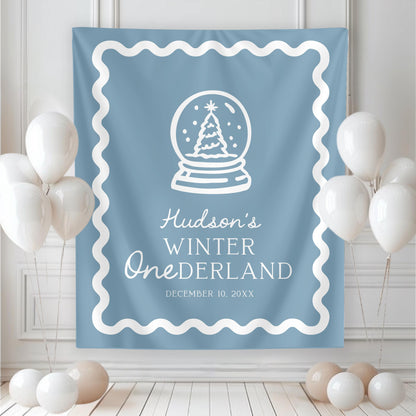 large backdrop hanging on wall.  blue with white graphics and wavy border.  christmas tree in snow globe and reads "hudson's winter ONEderland" with date of party below.  white balloons rest near both sides of banner