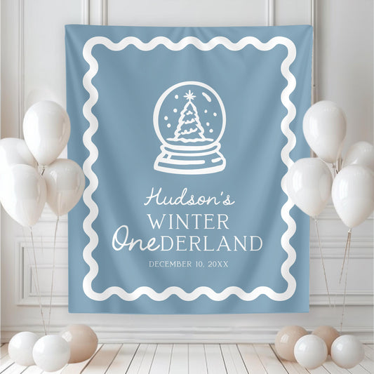 large backdrop hanging on wall.  blue with white graphics and wavy border.  christmas tree in snow globe and reads &quot;hudson&#39;s winter ONEderland&quot; with date of party below.  white balloons rest near both sides of banner