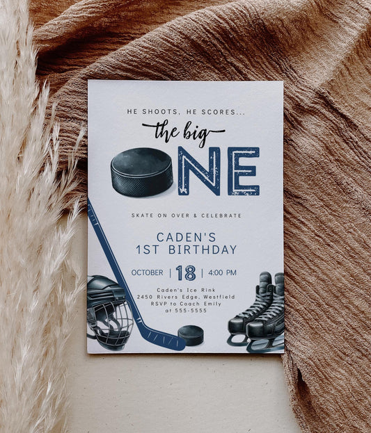 Hockey themed first birthday invitation with ‘he shoots he scores the big ONE’ text and mask, hockey stick, puck and skates graphics.