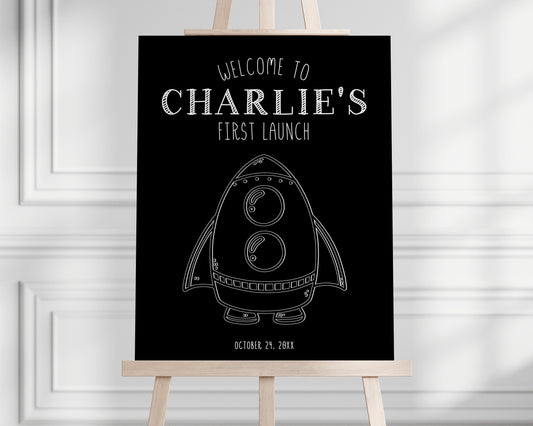 Black and white space party welcome sign. Black background with white graphics of rocket ship. Sign rests on an easel.