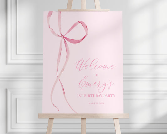 pink bow welcome sign resting on an easel.  pale pink background, minimalist design of pink bow graphic with long thin ribbons dangling alongside text.