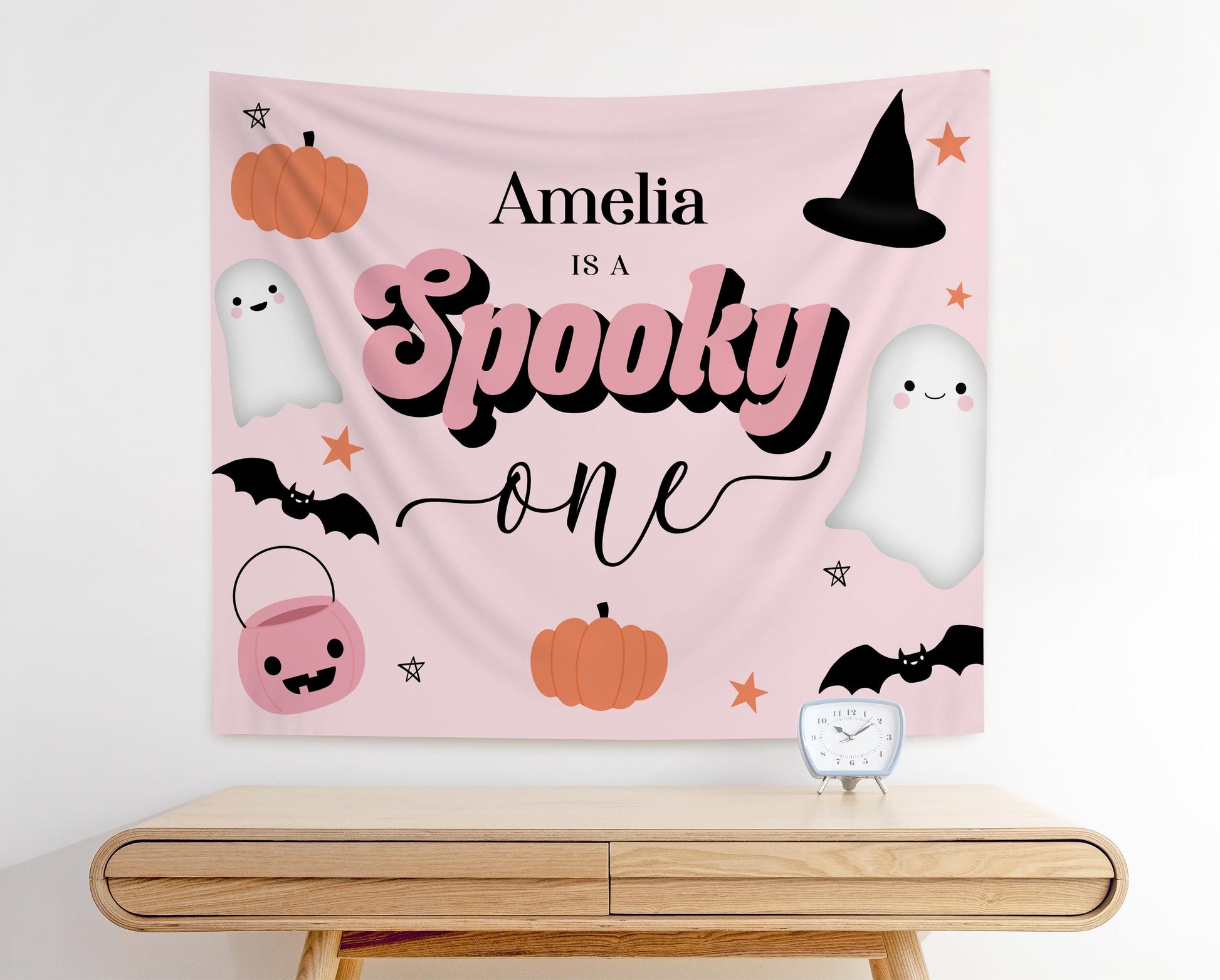 Pink Halloween first birthday backdrop with text “Amelia is a Spooky One,” featuring cute ghosts, pumpkins, bats, witch hat, candy bucket, and stars for a girly spooky birthday party decoration.