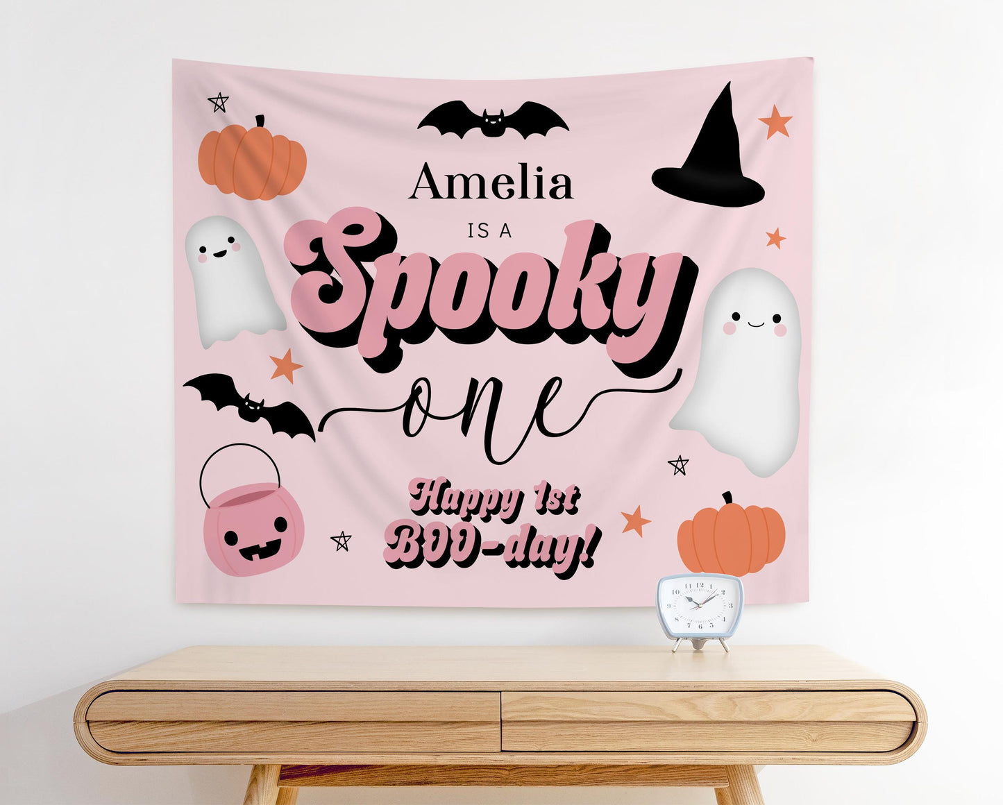 Pink Halloween first birthday backdrop with text “Amelia is a Spooky One, Happy 1st BOO-Day!” featuring cute ghosts, pumpkins, bats, witch hat, candy bucket, and stars for a girly spooky birthday party decoration.