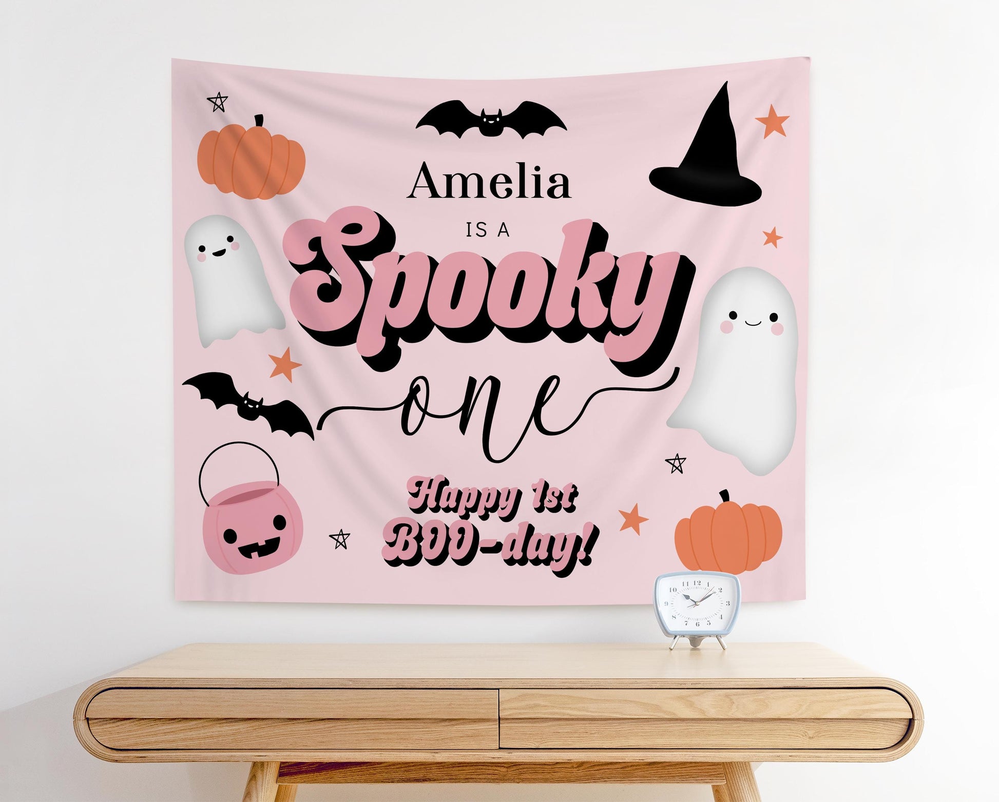 Pink Halloween first birthday backdrop with text “Amelia is a Spooky One, Happy 1st BOO-Day!” featuring cute ghosts, pumpkins, bats, witch hat, candy bucket, and stars for a girly spooky birthday party decoration.