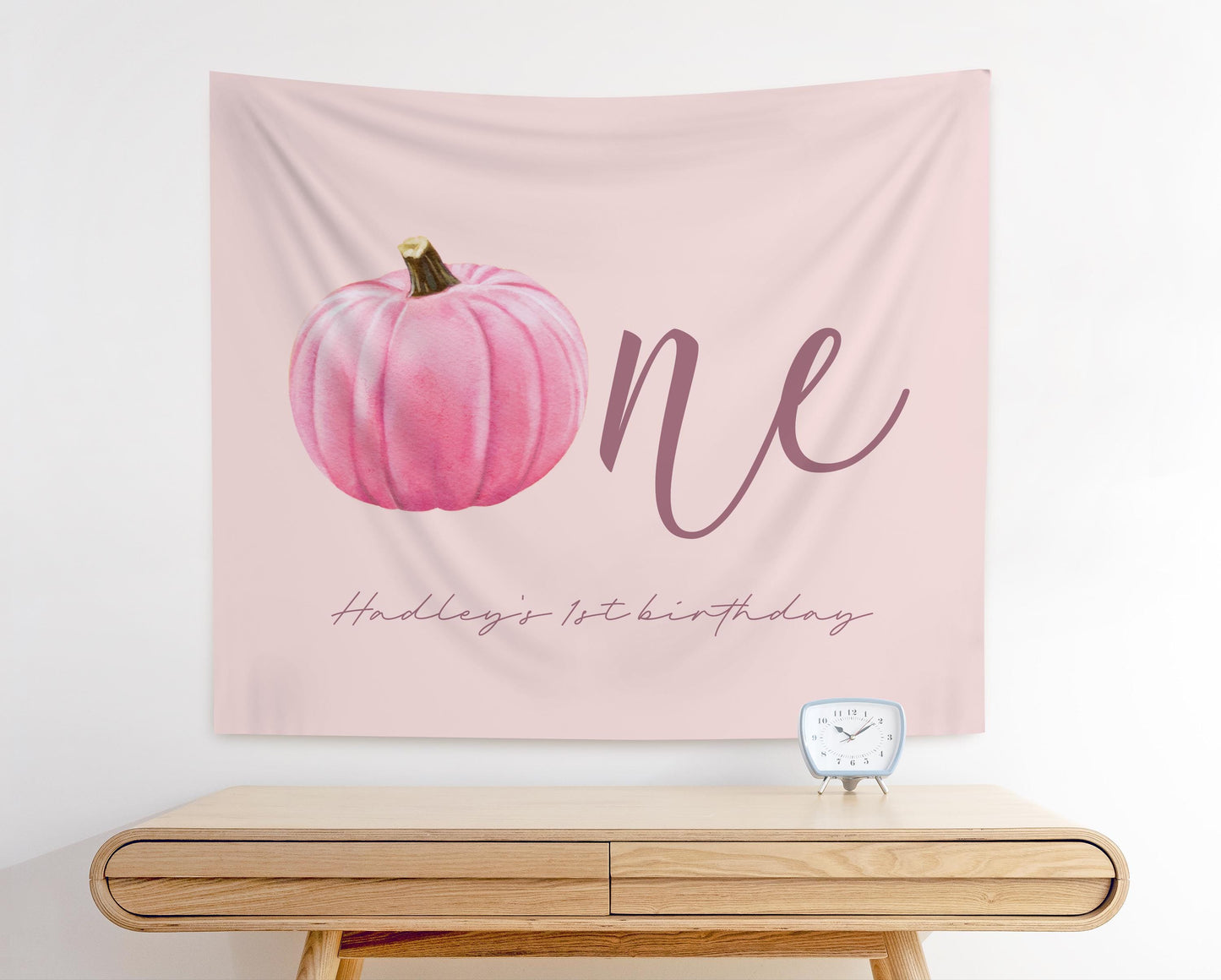 fabric backdrop with light pink background and pink pumpkin watercolor graphic. reads "ONE" with pumpkin as the "O".  Hadley's 1st Birthday written in script mauve color below.