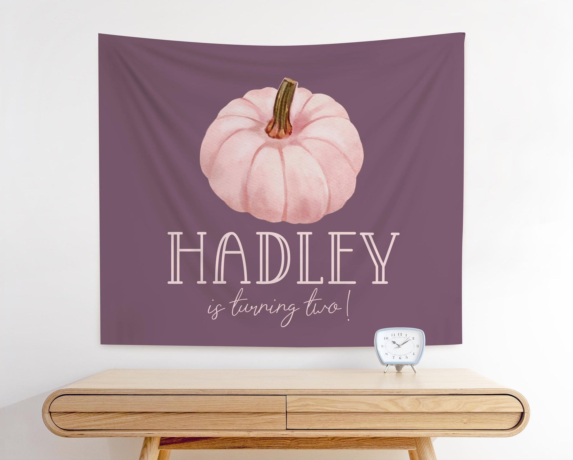 pink and purple fabric party backdrop for girl fall birthday party.  mauve background with pale pink watercolor pumpkin graphic.  Reads "Hadley is turning two".  Girl name in playful fun font and line below in modern handwritten font.