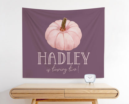 pink and purple fabric party backdrop for girl fall birthday party.  mauve background with pale pink watercolor pumpkin graphic.  Reads "Hadley is turning two".  Girl name in playful fun font and line below in modern handwritten font.
