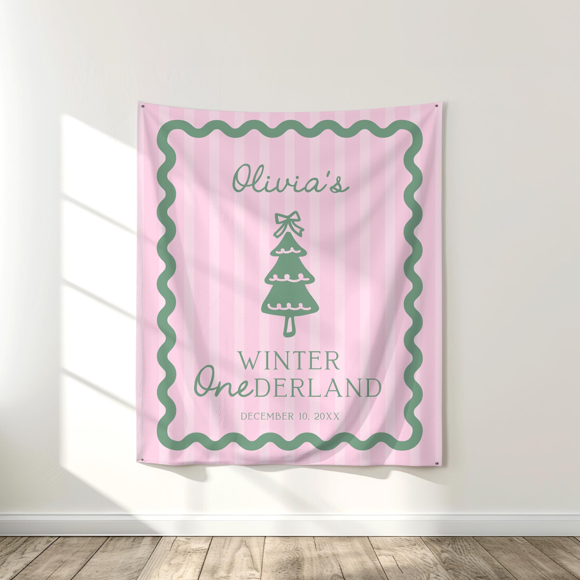large fabric backdrop hanging on white wall.  pink stripes and sage green wavy border.  Christmas tree graphic with bow on top, reads "Olivia's winter ONEderland" and date below.
