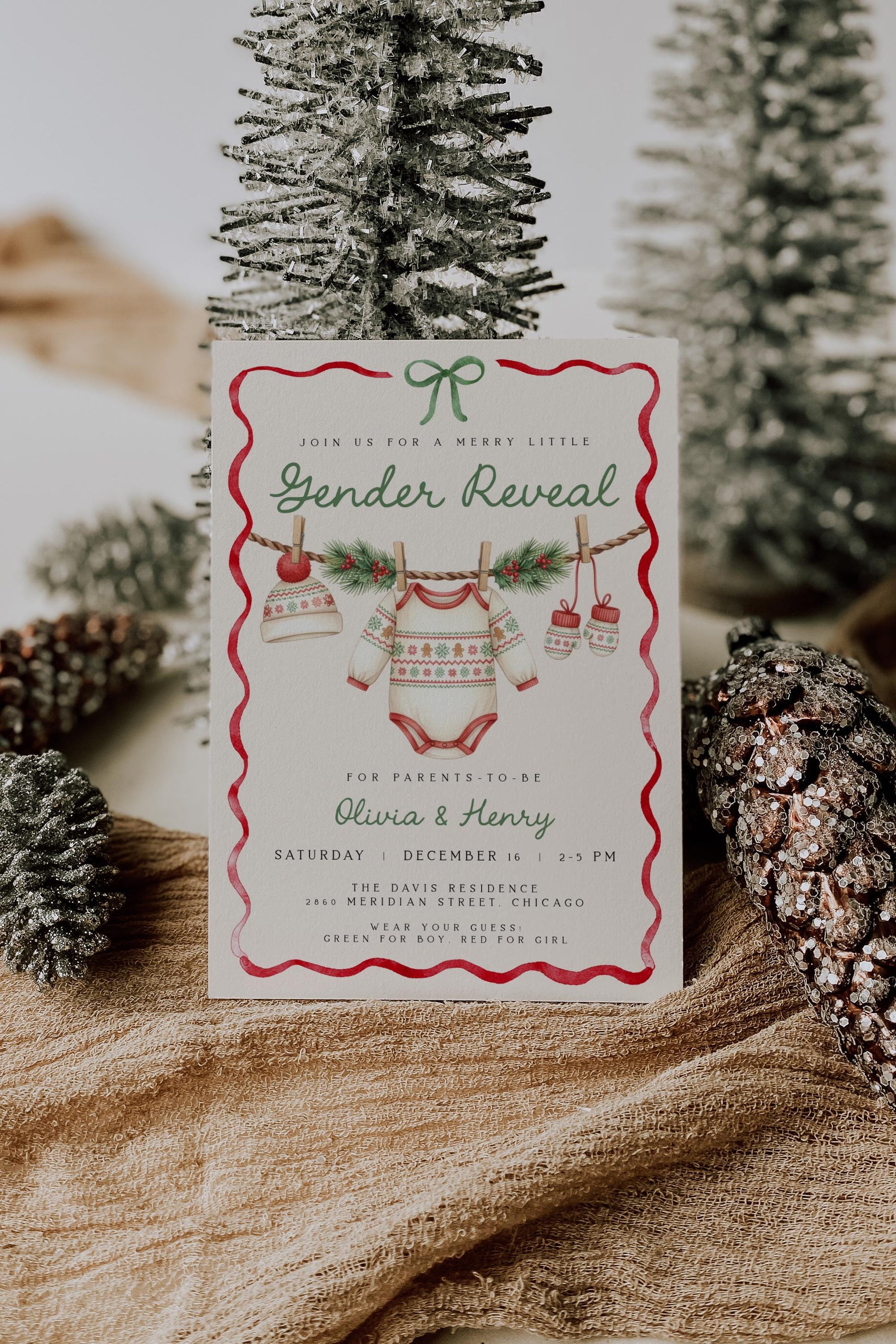 whimsical christmas gender reveal invitation featuring baby clothesline of winter onesie, hat and mittens.  watercolor red ribbon border with green bow.
