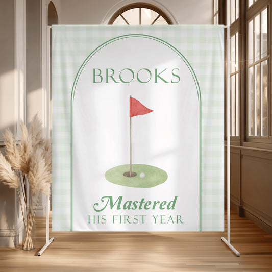 Personalized Hole in One first birthday backdrop with sage green gingham border, arch, watercolor golf flag and “Mastered His First Year” design.