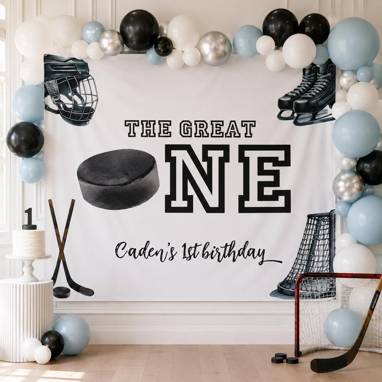 Personalized “The Great ONE” hockey first birthday backdrop featuring Caden’s name, displayed with blue, black, silver and white balloons, a birthday cake, hockey net, sticks and pucks.